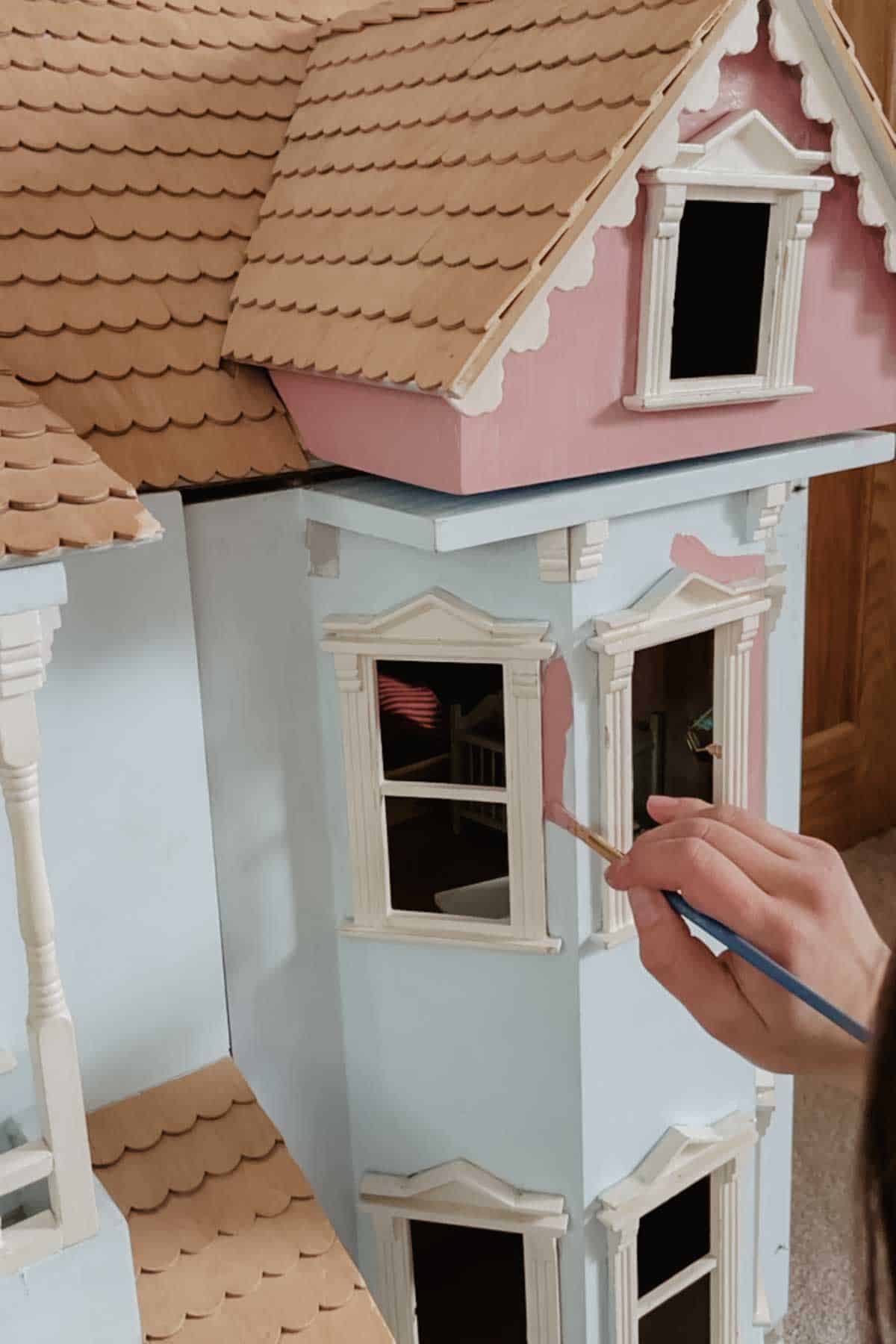 A lady paints a dollhouse with a muted pink paint and a small brush, cutting in around the windows. 