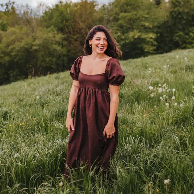 Elegant woman in a vintage-style maroon dress standing in lush green meadow with wildflowers and trees in background.