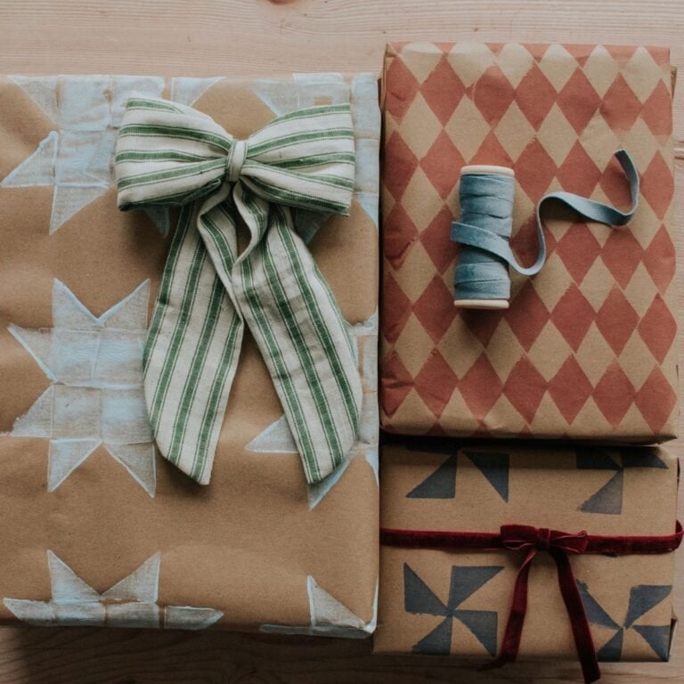Three gifts wrapped in brown potato stamp wrapping paper with stars, red diamonds, and geometric shapes. One has a green striped bow, another a red ribbon, and the third is topped with a spool of blue ribbon.