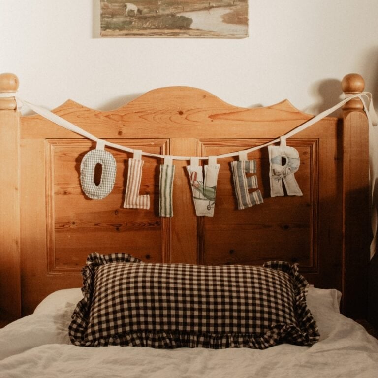 A wooden bed with a checkered pillow and a fabric banner spelling OLIVER across the headboard. A painting is mounted on the wall above the bed.