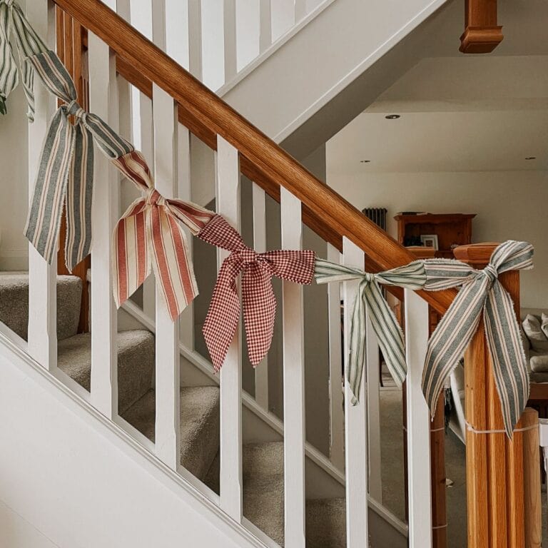 A staircase with a wooden handrail is decorated with a DIY Bow Garland—large fabric bows in stripes and checks tied along the railing. This no-sew Christmas decor adds a cozy, festive feel to the indoor space.