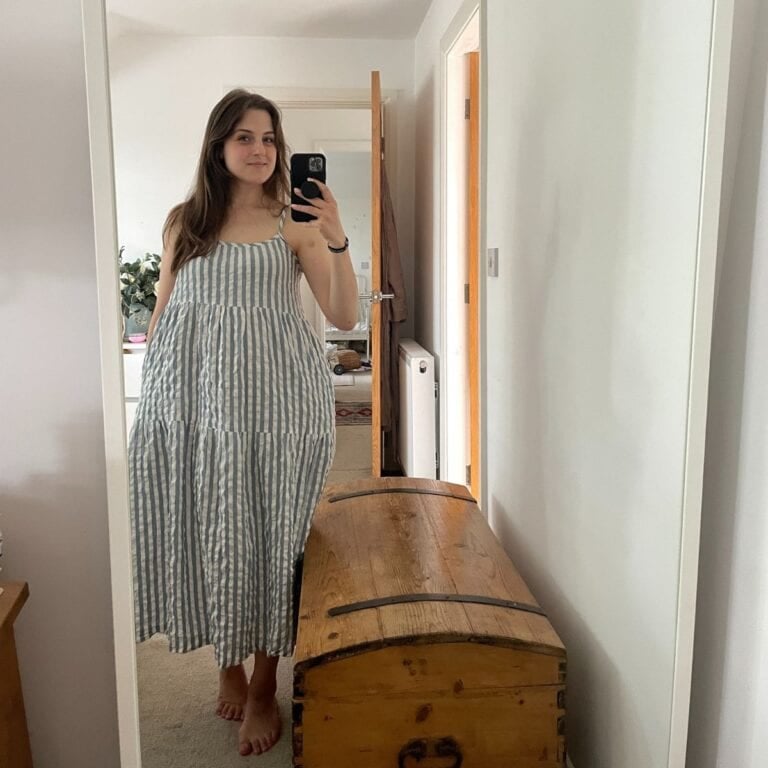 A woman in a blue and white striped dress, made from beginner sewing patterns, takes a mirror selfie in a bright room with light walls, a wooden trunk, and an open door in the background.