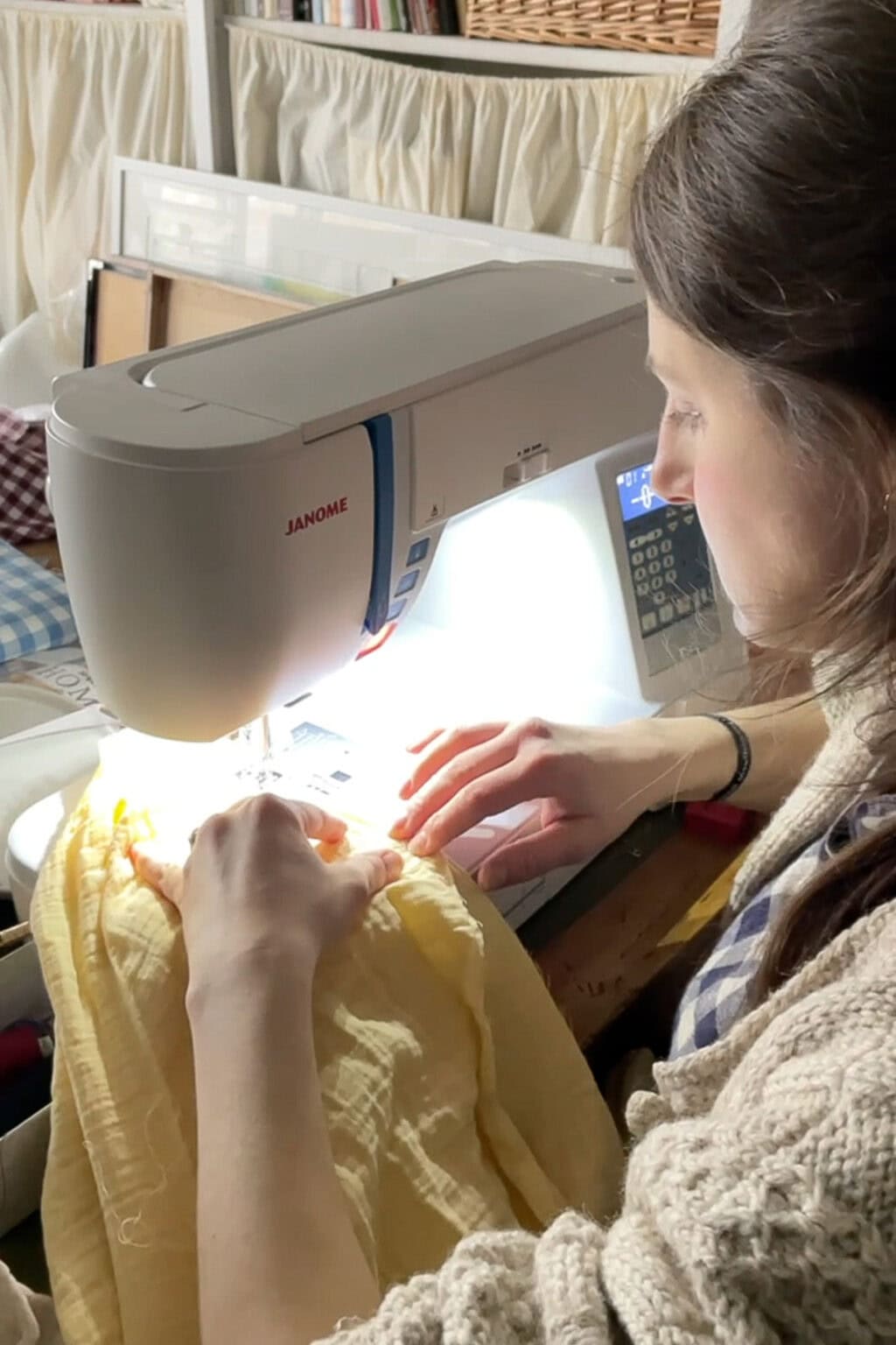 A woman sits at a sewing machine, guiding a piece of yellow fabric under the needle. She is focused on her work and surrounded by sewing materials in a cosy, well-lit room.
