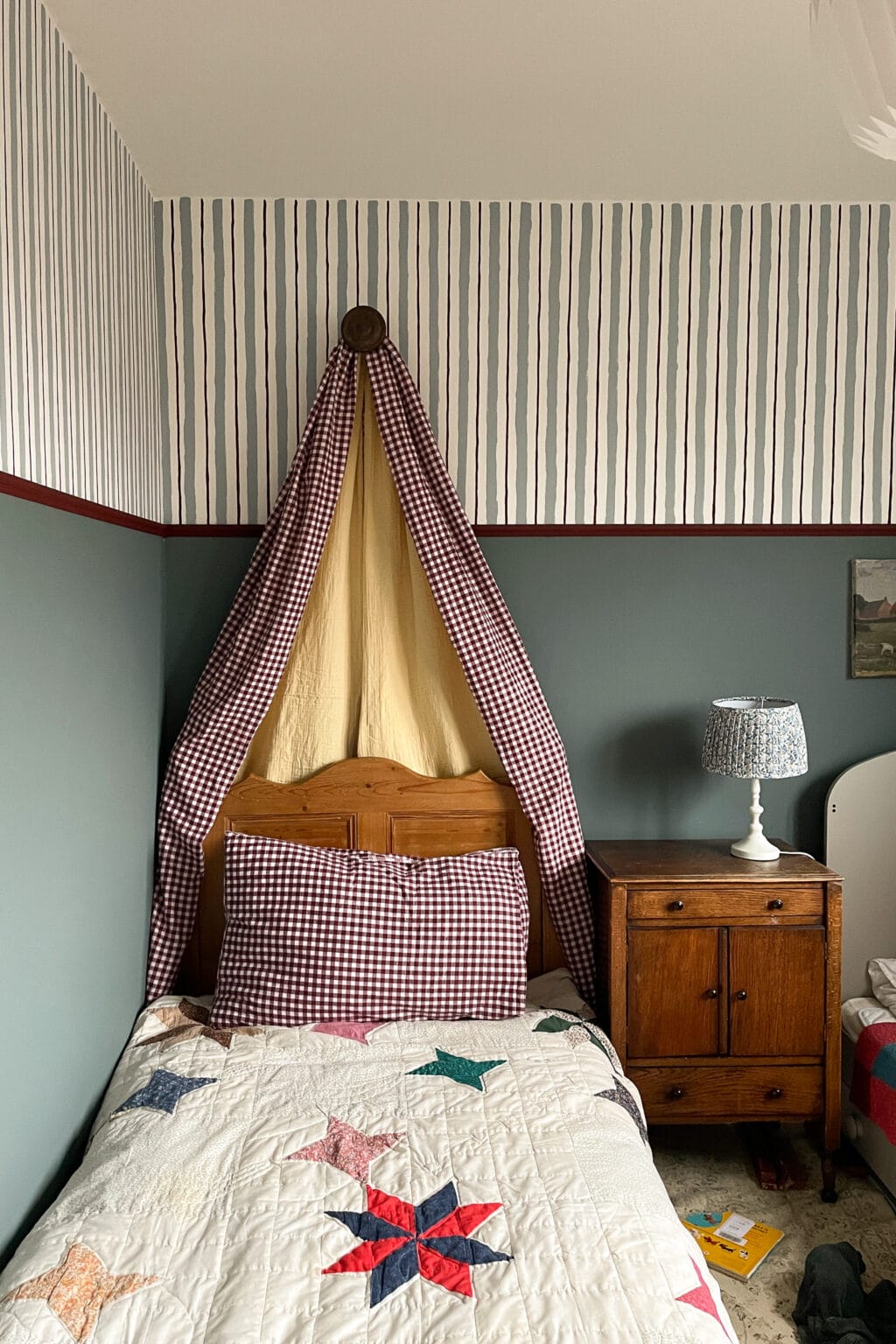 A cosy bedroom with a single bed featuring a star-patterned quilt, a large checked pillow, and a gingham canopy. Next to the bed is a wooden bedside table with a lamp and a small painting on the wall above.