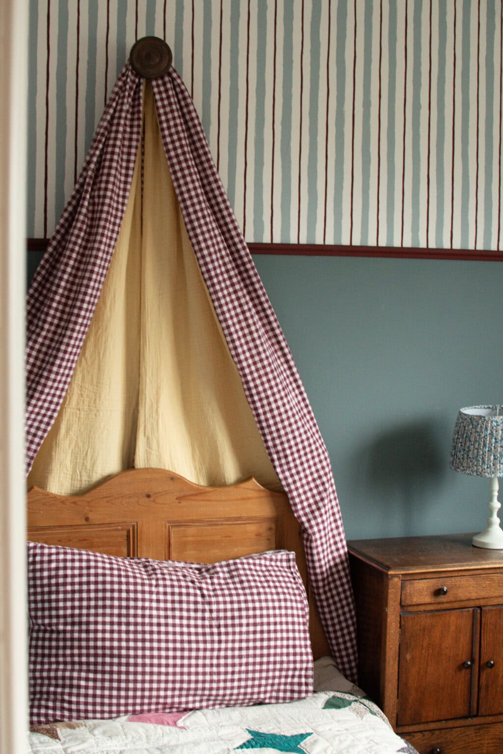 A cosy bedroom corner with a wooden bed, checked red-and-white pillow and curtains, pale yellow canopy, dark wood bedside table, and a lamp with a blue patterned shade, against blue and striped wallpapered walls.