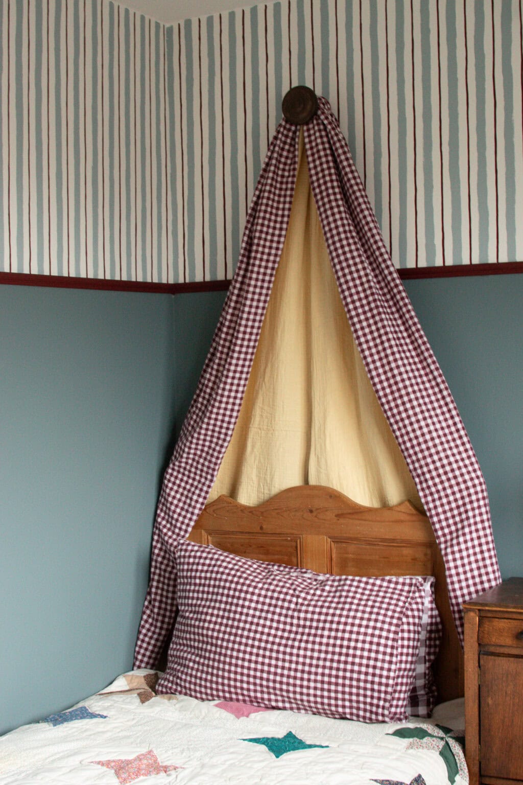 A cosy bedroom corner with a wooden bed, red and white chequered bedding, and a matching chequered canopy draped above the headboard. Walls are painted teal with a striped wallpaper border along the top.