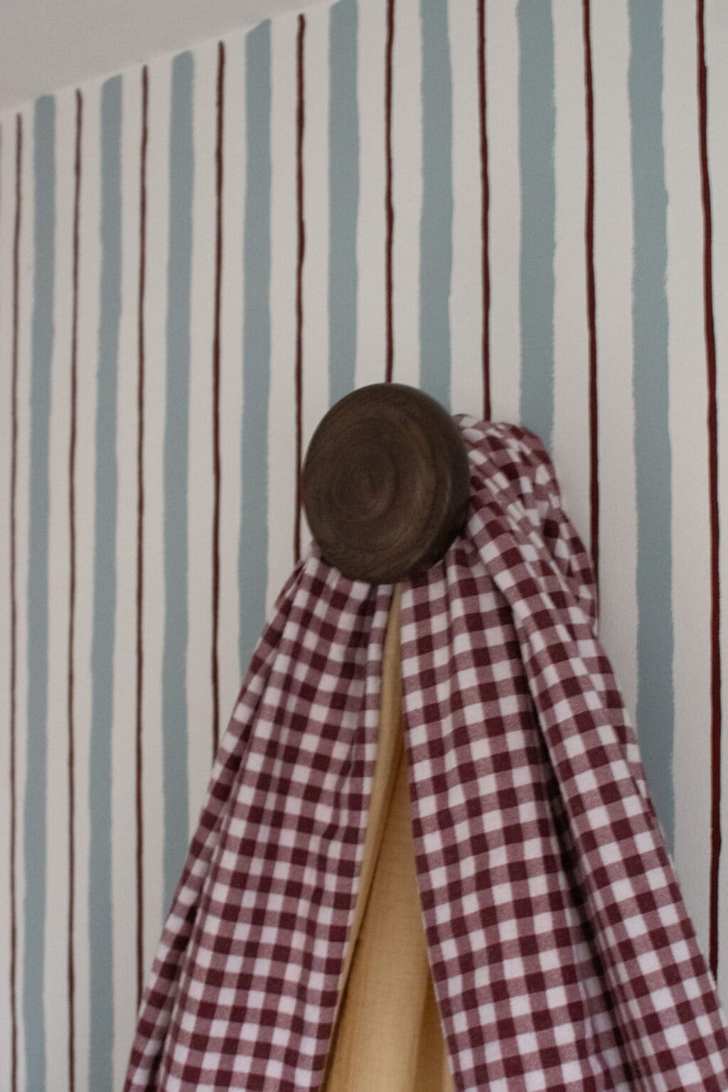 A brown wooden hook holds a red and white chequered cloth and a beige cloth against a wall with vertical blue and brown stripes.
