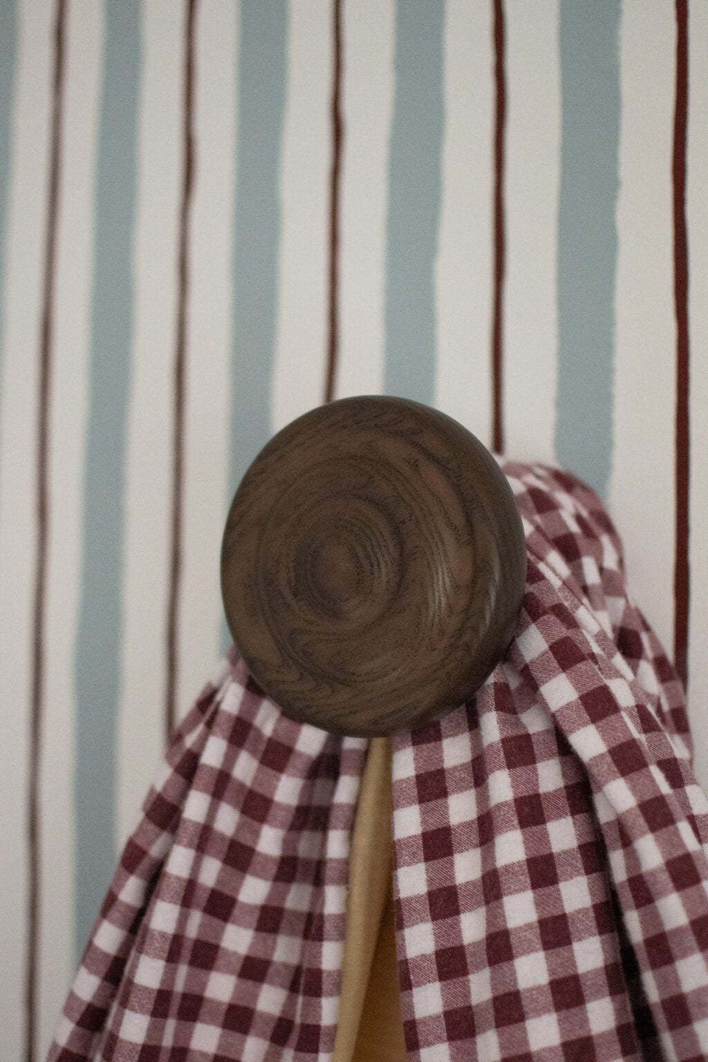A wooden wall hook holds a red and white chequered cloth, with a background of blue and white vertical striped wallpaper featuring thin brown lines.
