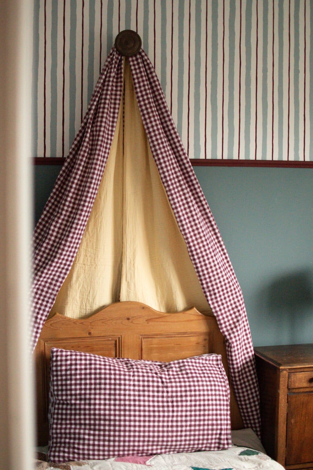 A wooden bed with a red and white chequered pillow and canopy, set against a wall with striped wallpaper and blue panelling, next to a wooden bedside table.