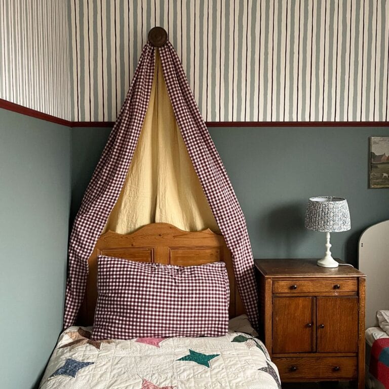 A small bed with a wooden headboard, checked red and white pillow and canopy, yellow fabric behind, patchwork quilt, wooden bedside table with a lamp, and striped wallpaper above green walls.