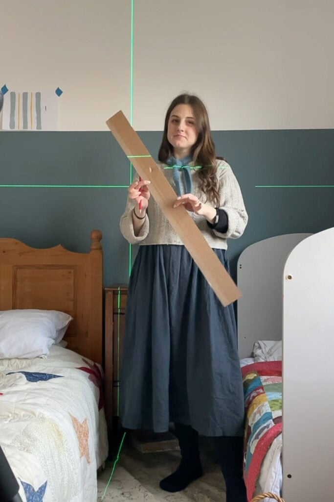 A woman stands between two beds in a bedroom, holding a long piece of cardboard. Green laser lines are projected on the wall and furniture, likely to guide alignment or assist in painting wall stripes with precision.
