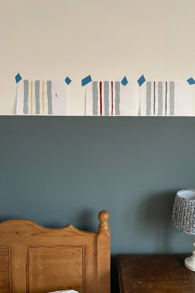 Sheets of paper with different paint swatches taped to a white wall above a blue-grey feature wall, perfect for planning or painting wall stripes, with part of a wooden headboard, brown bedside table, and lamp visible below.