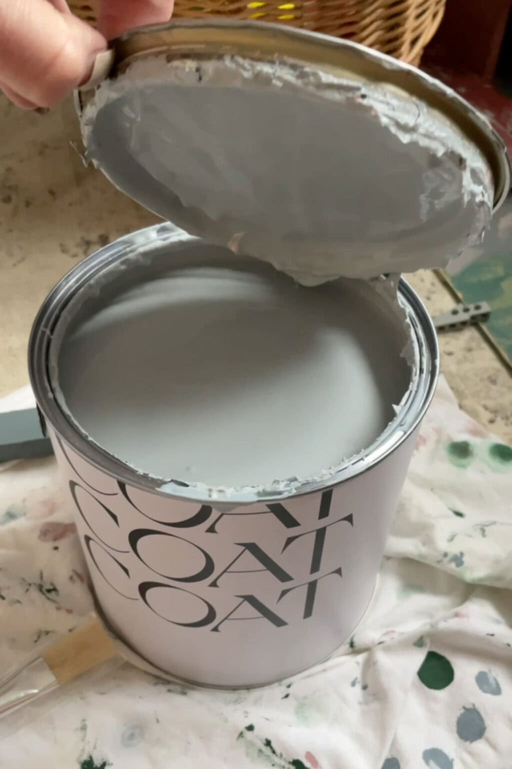 A hand opens a tin of light grey paint labelled COAT on a paint-splattered cloth, with a paint stirrer partially visible nearby.