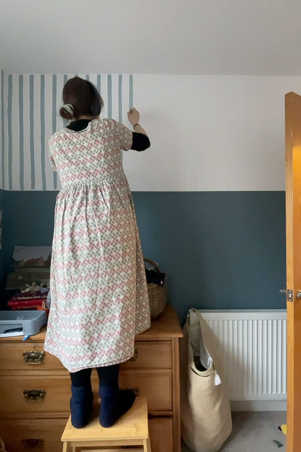 A woman in a patterned dress stands on a wooden stool, painting stripes—vertical blue lines—on the upper part of a white wall in a room with blue panelling and wooden furniture.