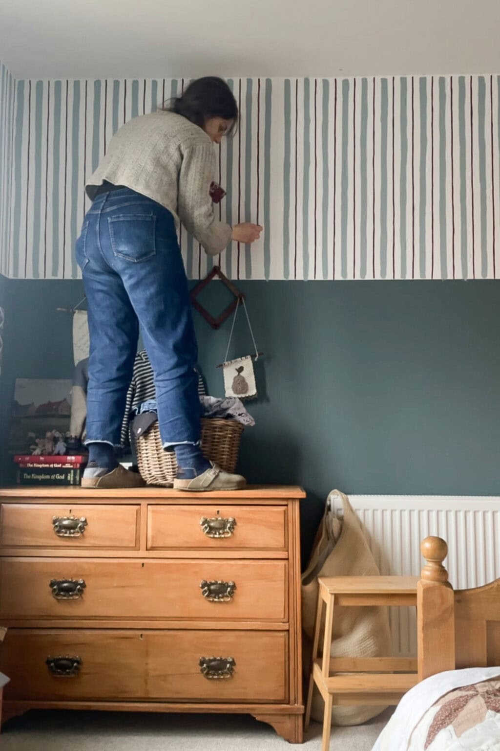 A woman stands on top of a wooden chest of drawers, painting red stripes on the wall of a bedroom. She reaches up with care, decorating the space whilst furniture and decor items surround her.