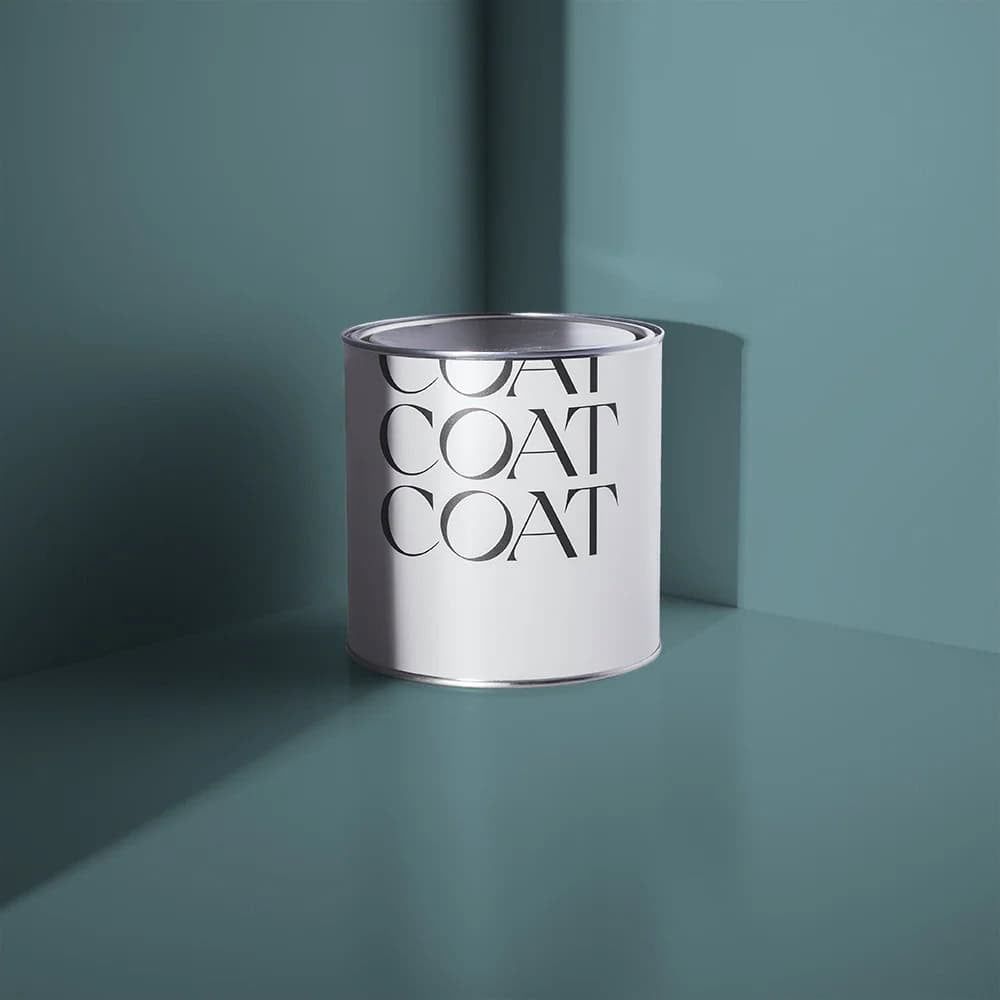 A white paint tin labelled COAT three times in bold black letters sits on a teal surface against a matching teal wall—perfect for painting wall stripes—with soft light and shadows creating a minimalist look.
