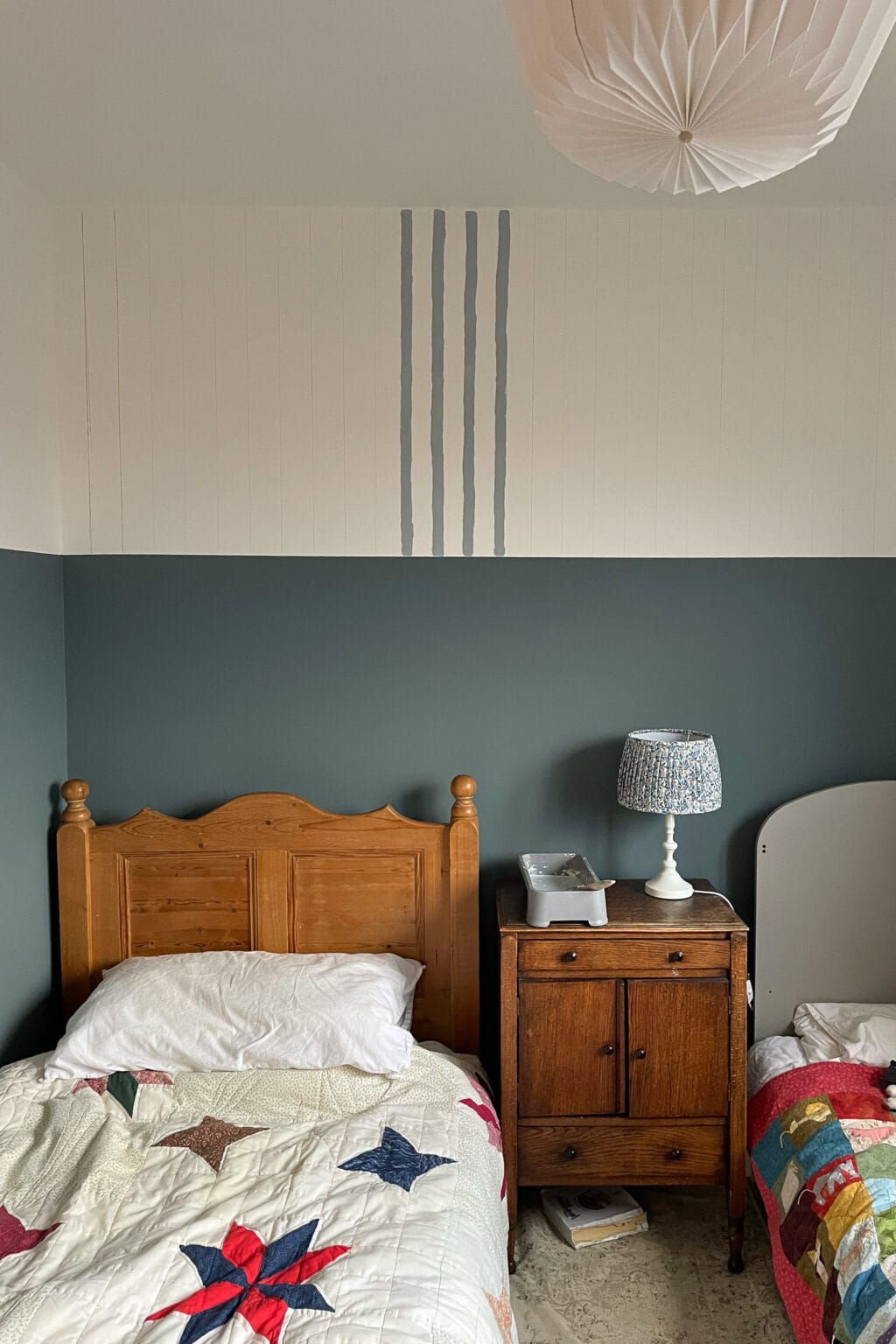 Two twin beds with star quilts sit beside a wooden bedside table with a blue patterned lamp and a telephone. The walls are half dark green and half white, with four painted wall stripes in blue grey above the beds.