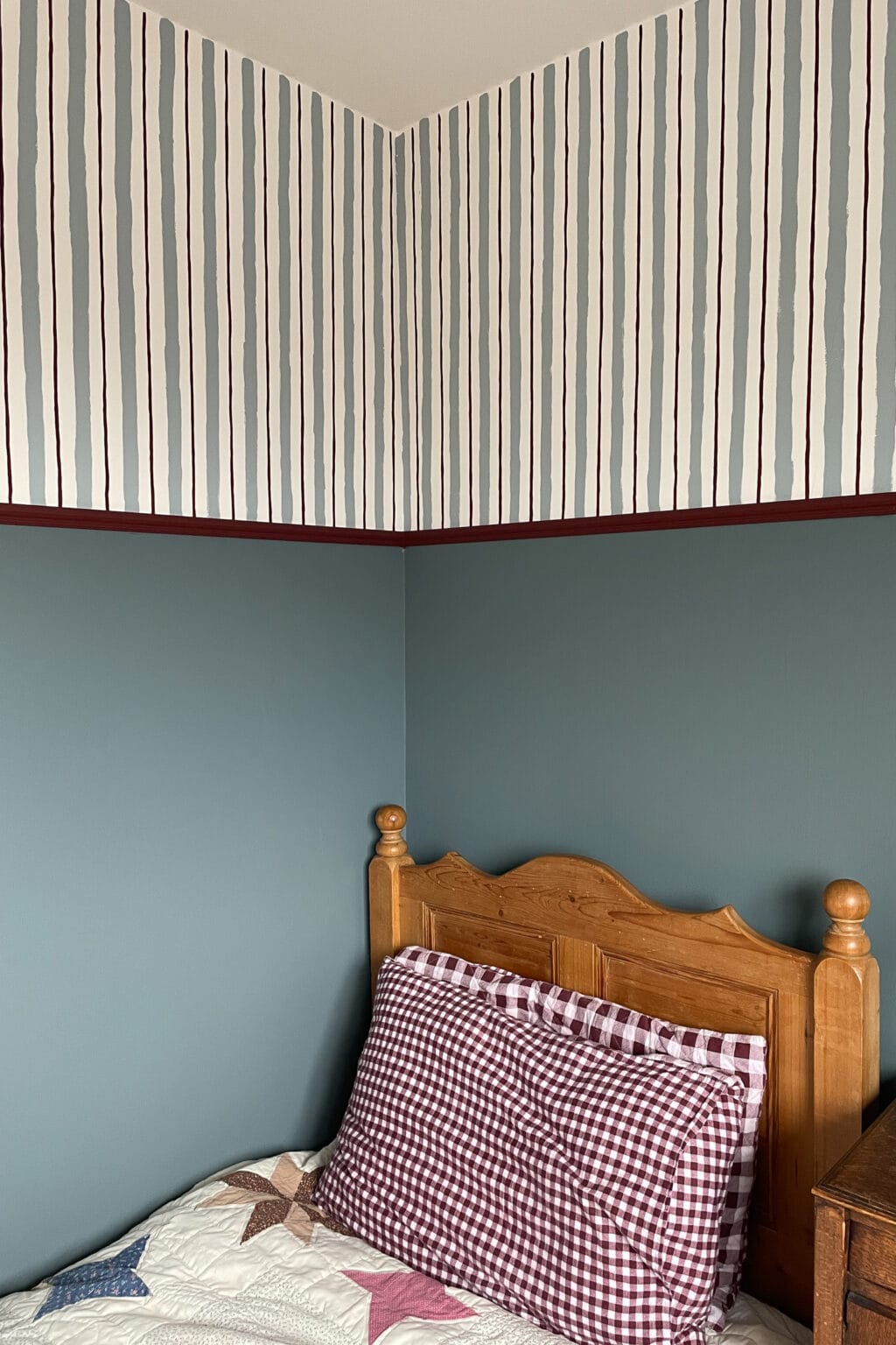 A cosy bedroom corner with teal walls, a wooden bed frame, a burgundy gingham cushion, a patchwork quilt, and striped wallpaper in burgundy, blue grey, and white above a rich burgundy trim.