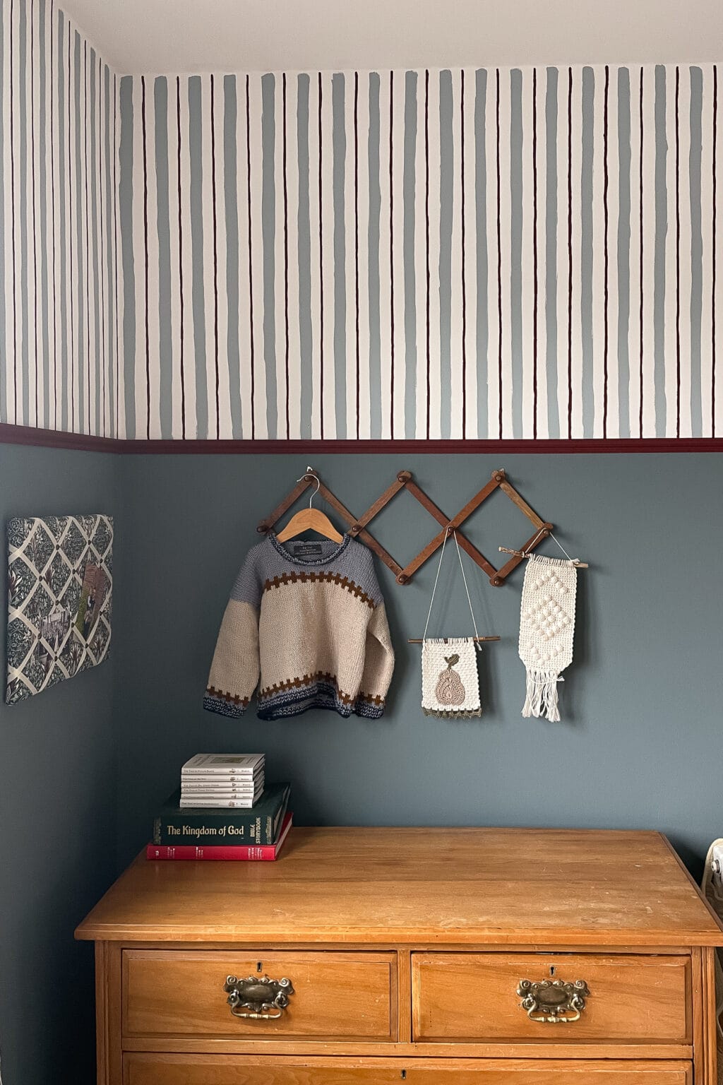 A wooden chest of drawers sits against a blue wall with a striped upper section, perfect for painting wall stripes. Above the chest of drawers, a wooden rack holds a knitted baby jumper and two crocheted wall hangings. Books are stacked neatly on the chest of drawers.