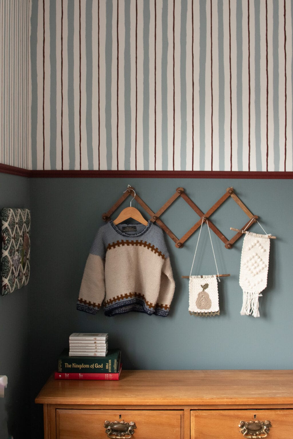 A cosy room with blue-green walls features a wooden chest of drawers, folded books, a child’s knitted jumper on a rail, and two pieces of macramé art. The upper wall is adorned with painted wall stripes for an extra touch of charm.