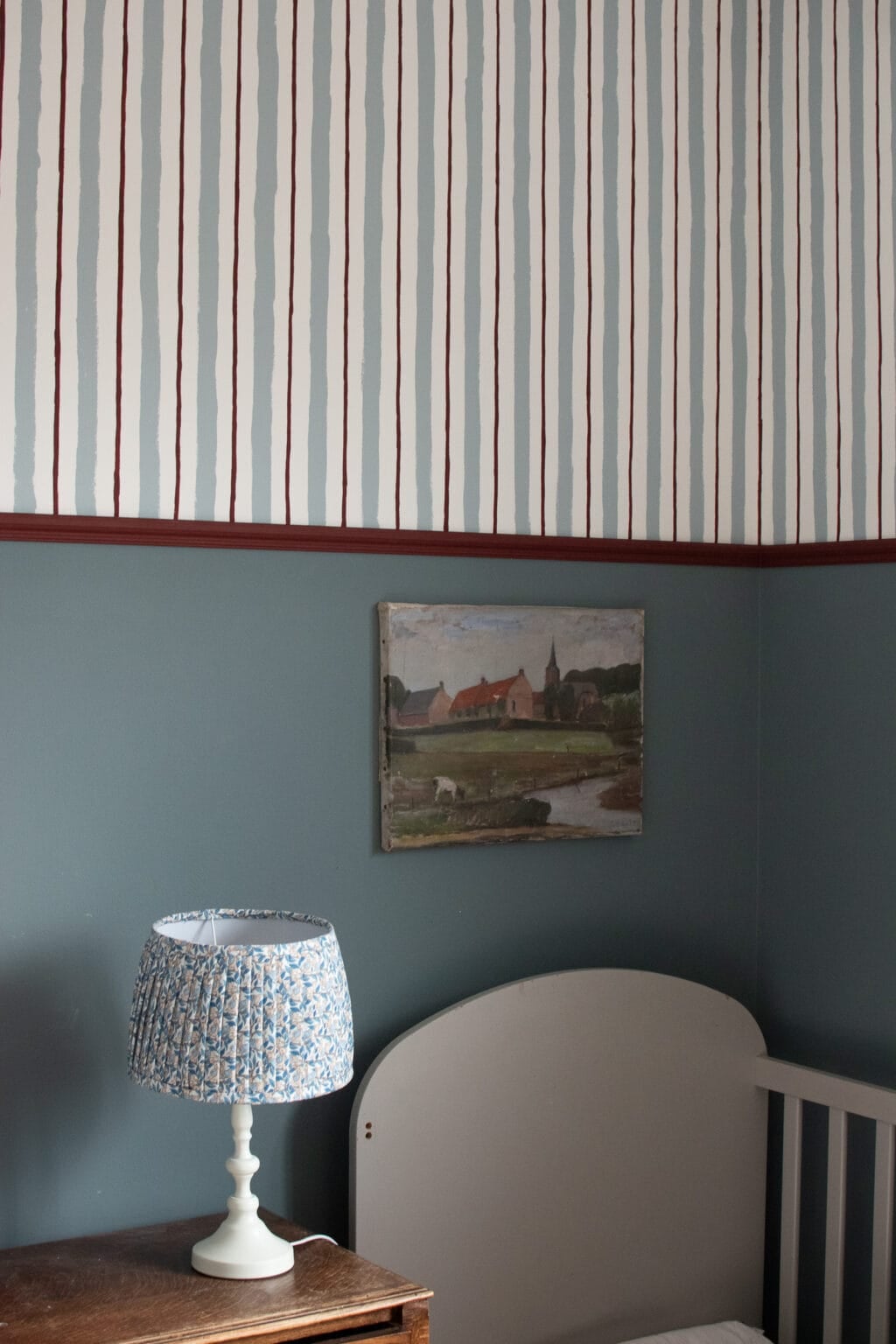 A cosy corner of a child's room with blue wall with the upper half painted with vertical red and blue stripes, a cot, a small wooden table, a floral patterned lamp, and a landscape painting on the wall.