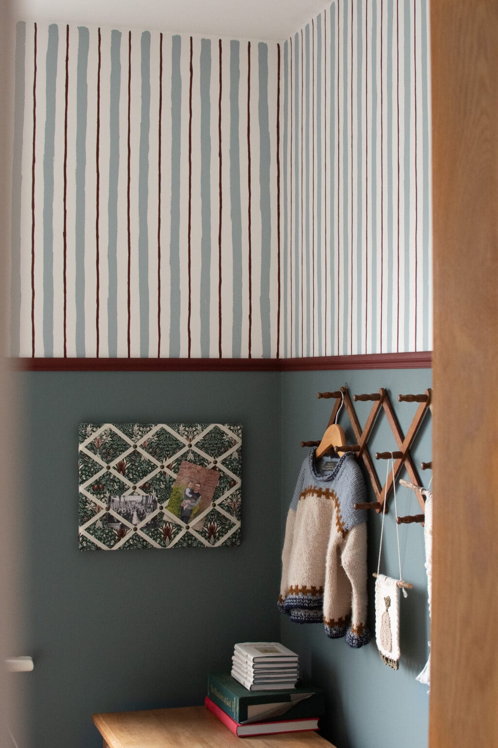 A small room with wall stripes painted above as vertical wallpaper, a wooden desk with stacked books, a wall-mounted rack holding two knitted jumpers, and a lattice noticeboard displaying photos.