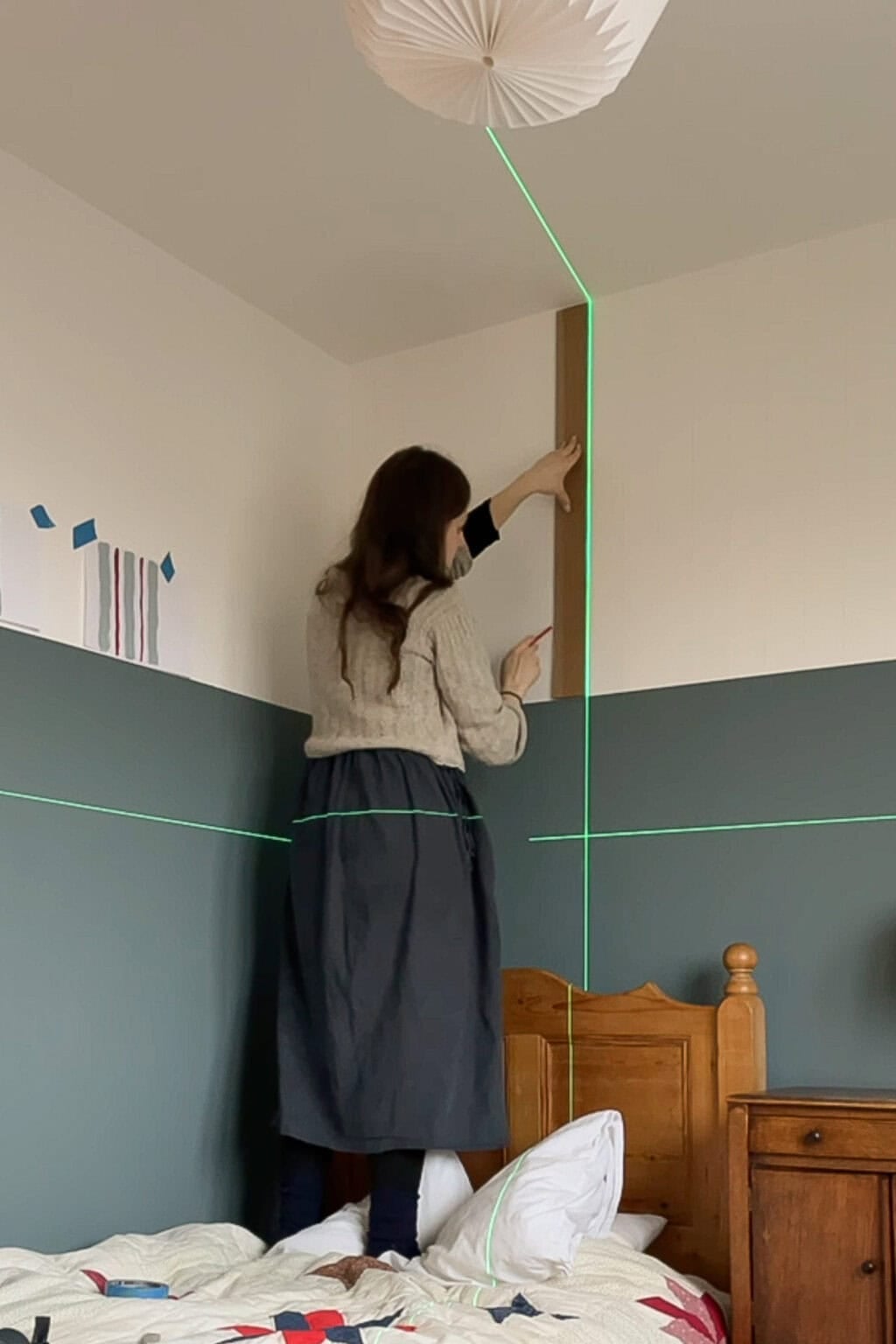 A woman stands on a bed, holding a wooden board against a wall split by a green laser level line. She aligns the board vertically near the room’s corner above a wooden headboard, preparing to start painting.
