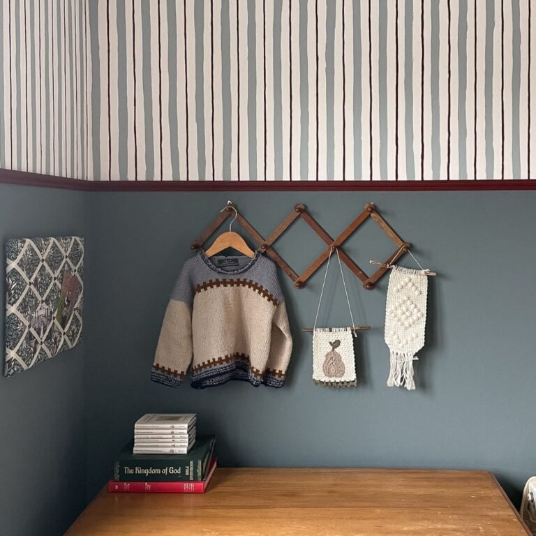 A children's jumper, two macramé wall hangings, and a few books sit on a wooden desk against a blue wall with striped wallpaper, reminiscent of painting wall stripes, and a foldable wooden wall rack.
