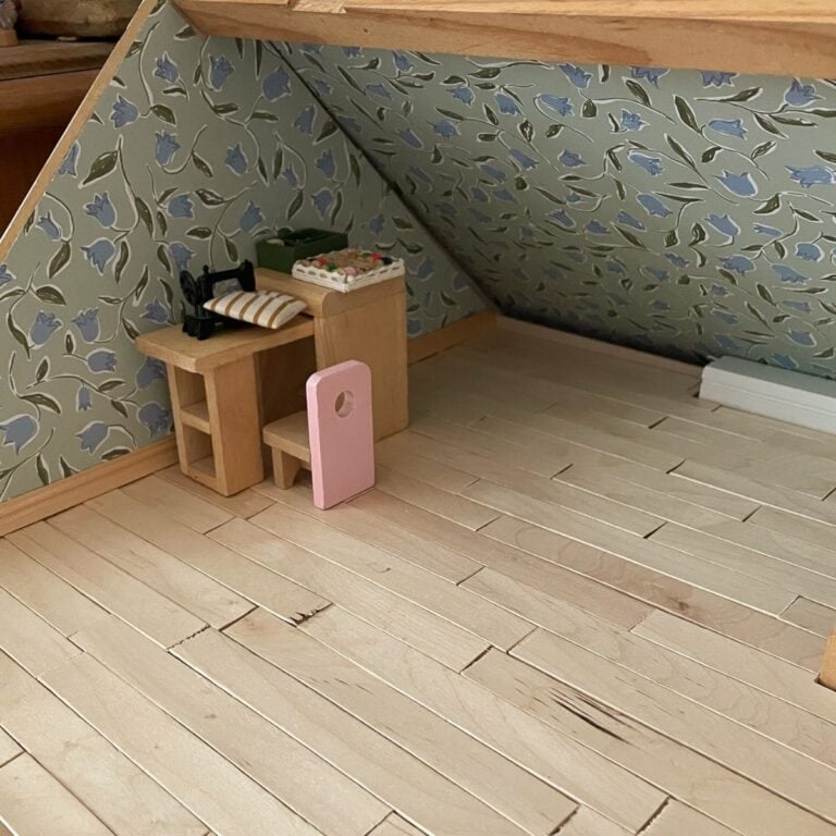 A miniature attic room with light wood floor, blue floral wallpaper, and a small wooden desk and chair in the corner. The desk holds a black sewing machine, fabric, and sewing supplies.