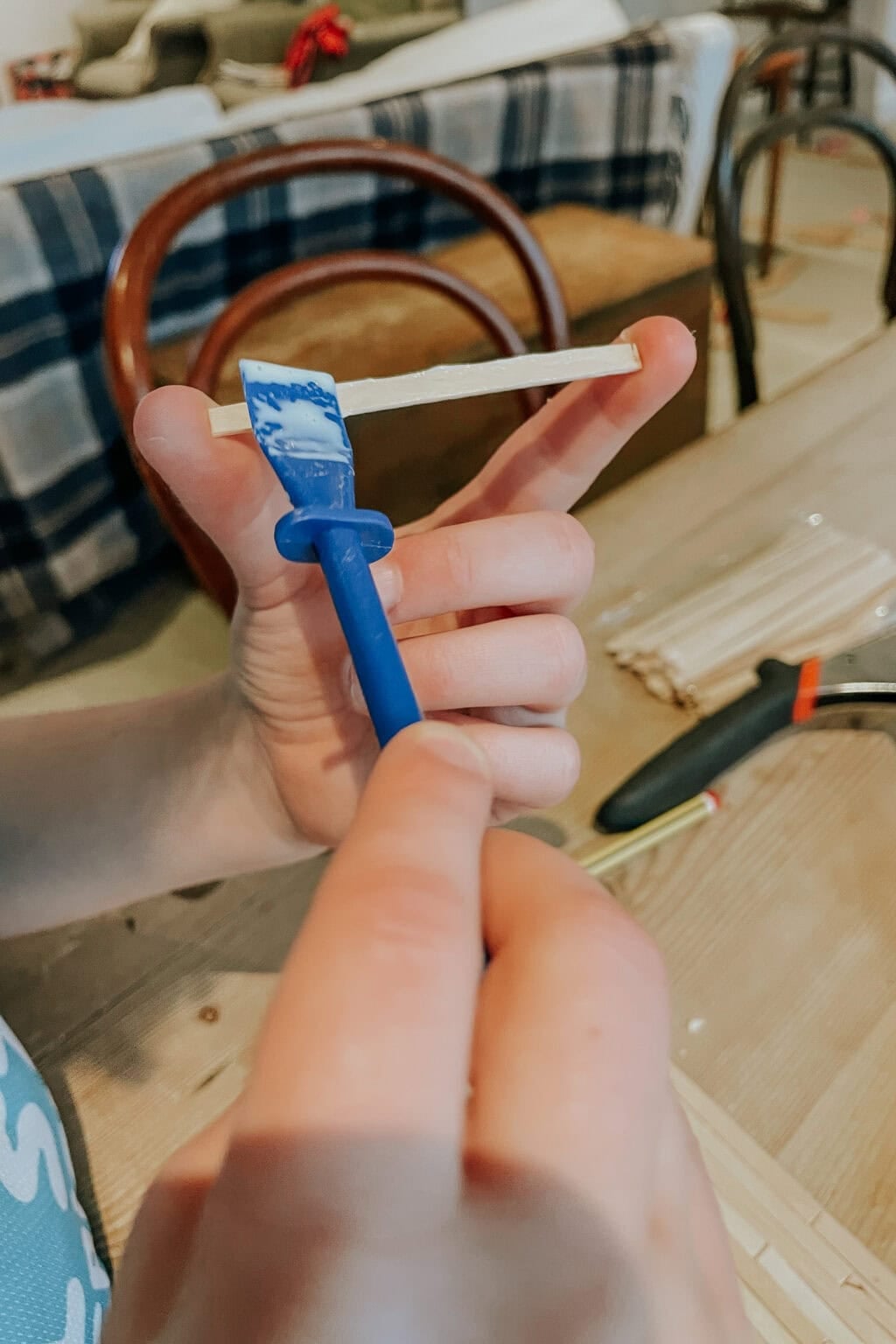 A person holds a blue plastic tool applying glue to the end of a wooden stick, with other craft supplies and a chequered blanket visible in the background.