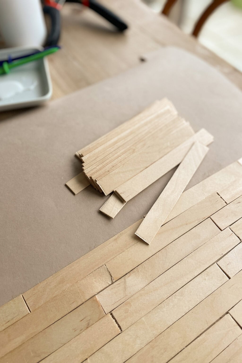 A pile of thin wooden sticks rests on a table covered with brown paper, next to a partially completed arrangement of wooden sticks laid out in rows, likely for a craft or model project.