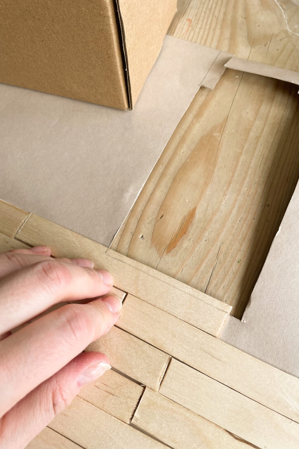 A hand is placing wooden sticks in a neat row on a wooden surface, next to a cardboard structure and a cut-out section of paper.