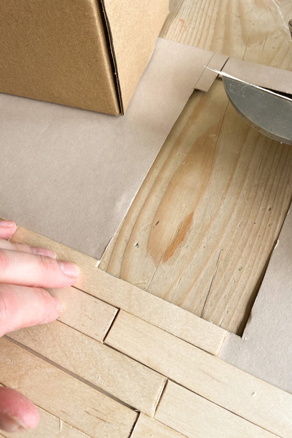 A hand presses wooden craft sticks onto a flat surface covered with paper, next to a rectangular hole revealing a wooden table underneath, with part of a cardboard box and a roll of tape visible.