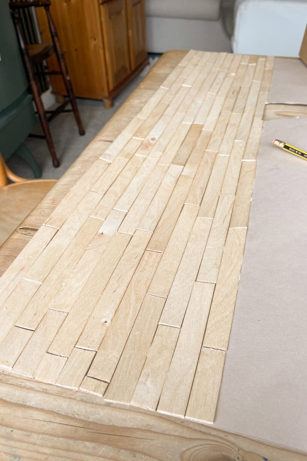 A wooden surface covered with small, rectangular wooden tiles arranged in a staggered pattern, to create a DIY dollhouse flooring. A pencil and some furniture are visible in the background.