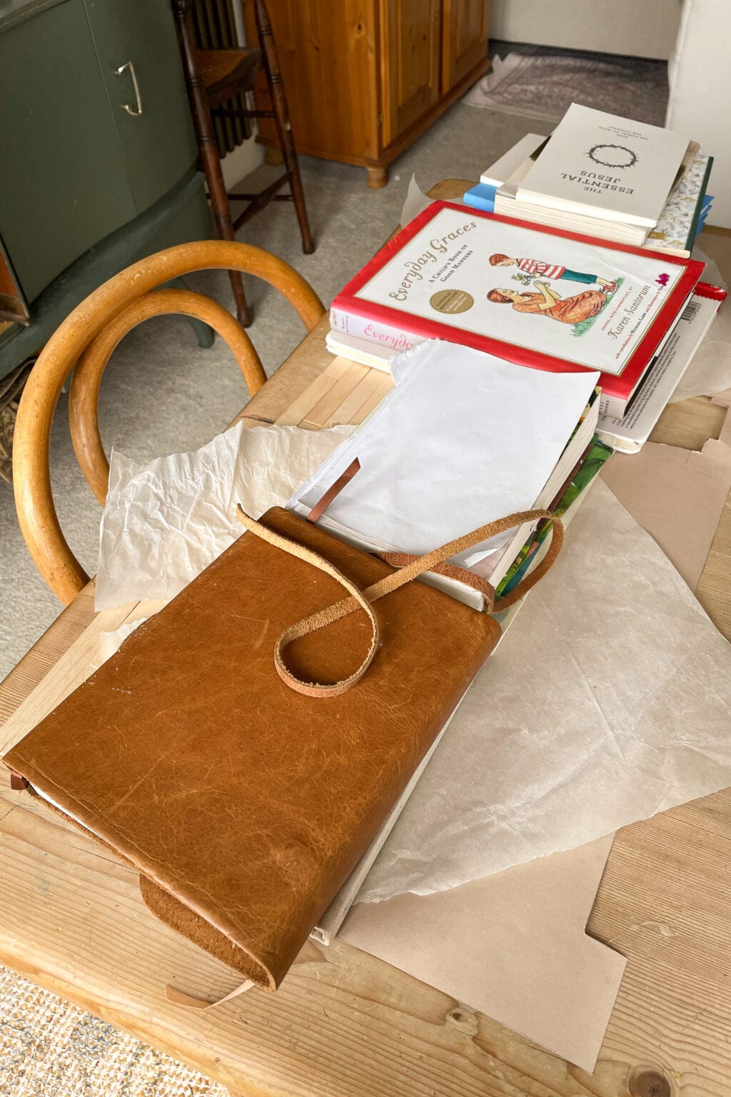 A tan leather diary with a strap lies open on a wooden table beside a stack of assorted books and tissue paper. Curved wooden chair backs and a wooden cupboard are visible in the background.