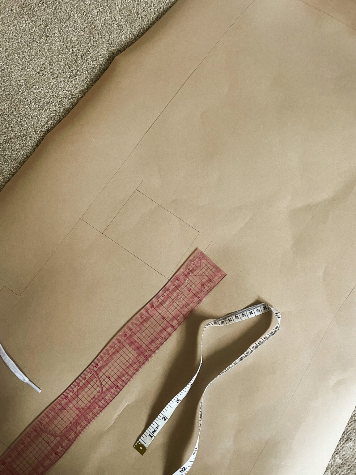 A large sheet of brown pattern paper with pencil markings, a transparent red ruler, a white measuring tape, and part of a white object, all on a carpeted floor.