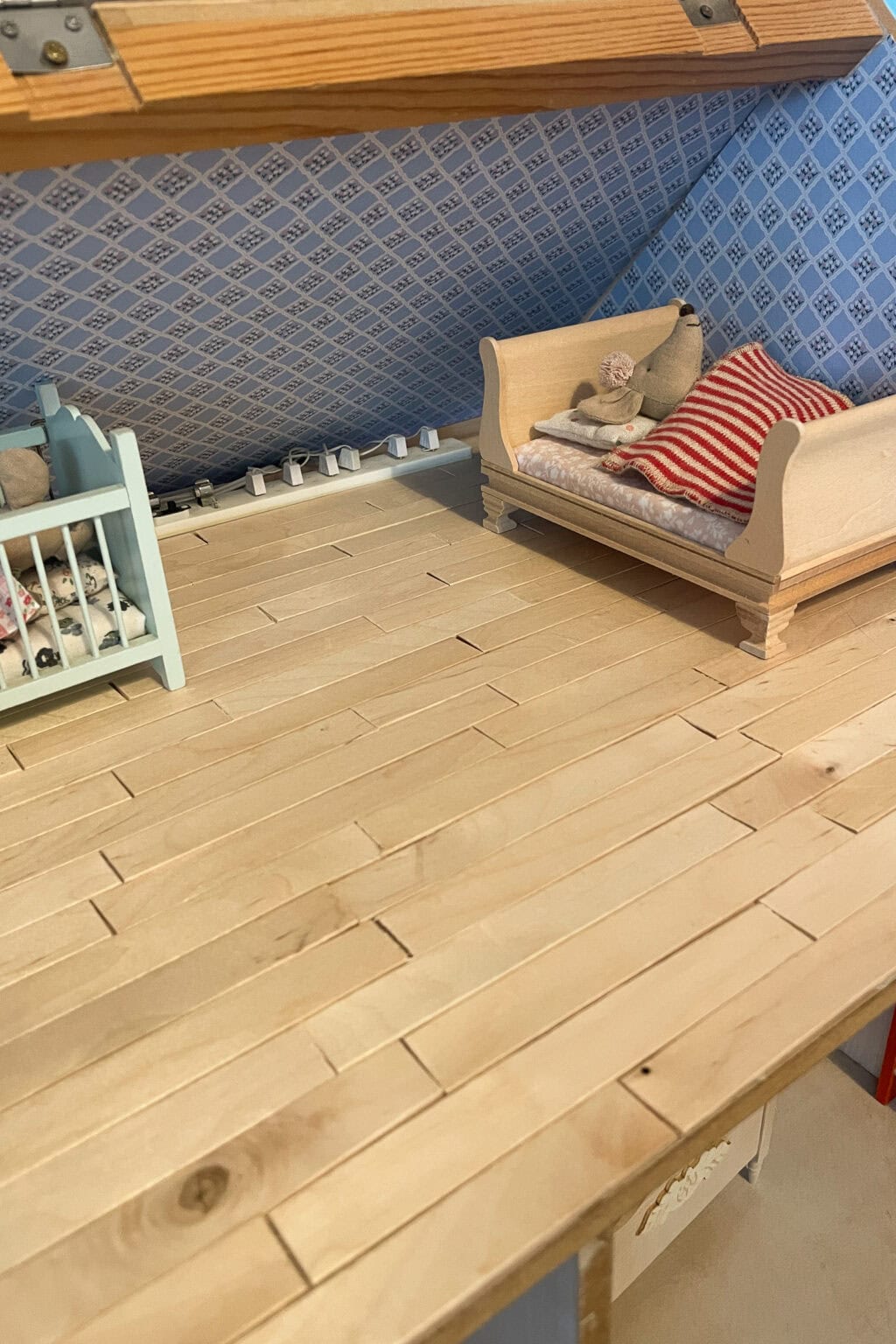 A dolls' house room with a DIY dollhouse flooring, a small blue cot with dolls, and a beige doll bed with a red and white striped blanket and a toy mouse, set against blue patterned wallpaper.