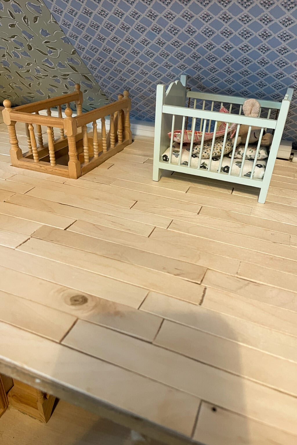Two miniature cots, one wooden and one white with bedding and a small toy, sit on a light wooden floor in a dolls' house room with patterned wallpaper.