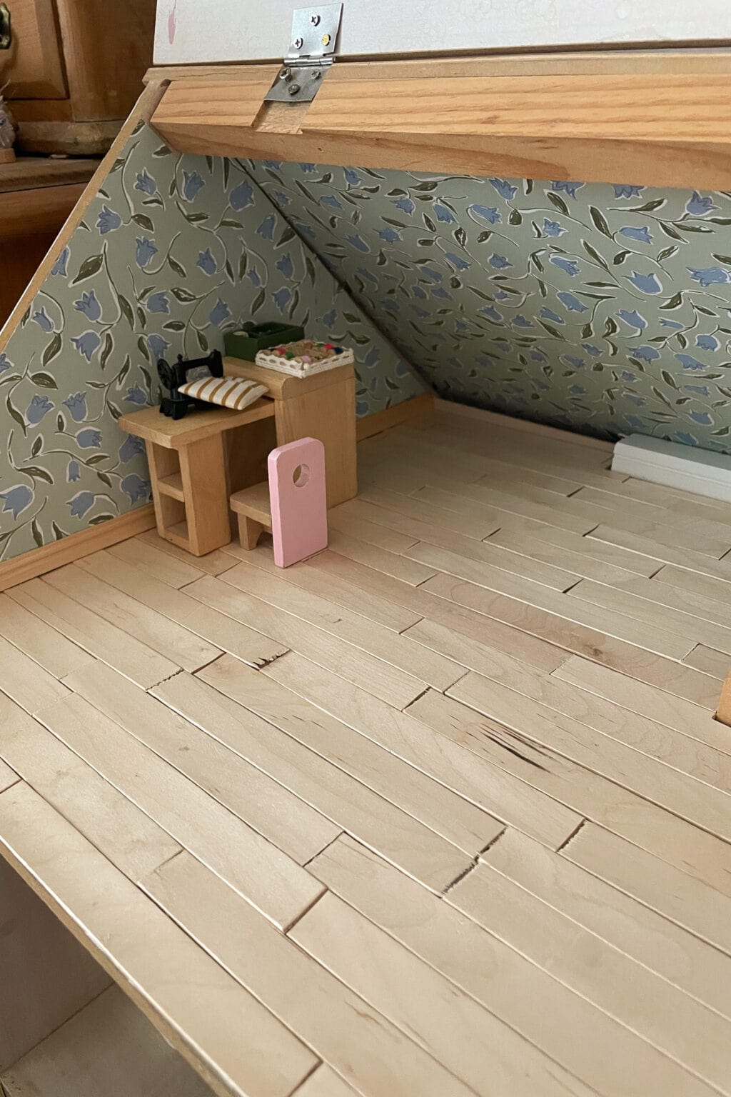 A miniature attic room with wooden plank flooring, wallpapered sloped walls, a small desk with items, a chair, and a pink door propped against the desk. The room is partially furnished.