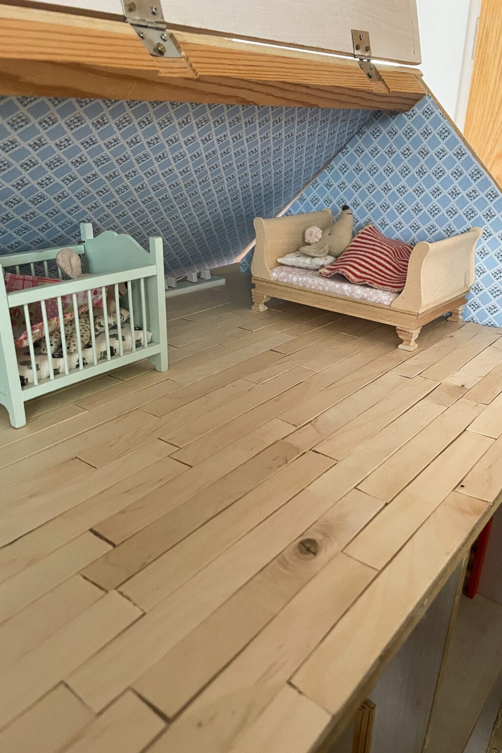 A miniature attic room with a wooden floor, featuring a small cot with blankets and a doll, and a bed with pillows and a teddy bear, against blue patterned wallpaper.