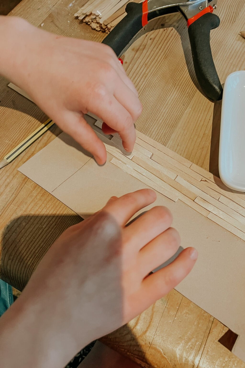 Two hands arrange wooden sticks on a piece of cardboard on a wooden table. Nearby are a pair of pliers and a small white dish. The scene suggests a craft or DIY project in progress.