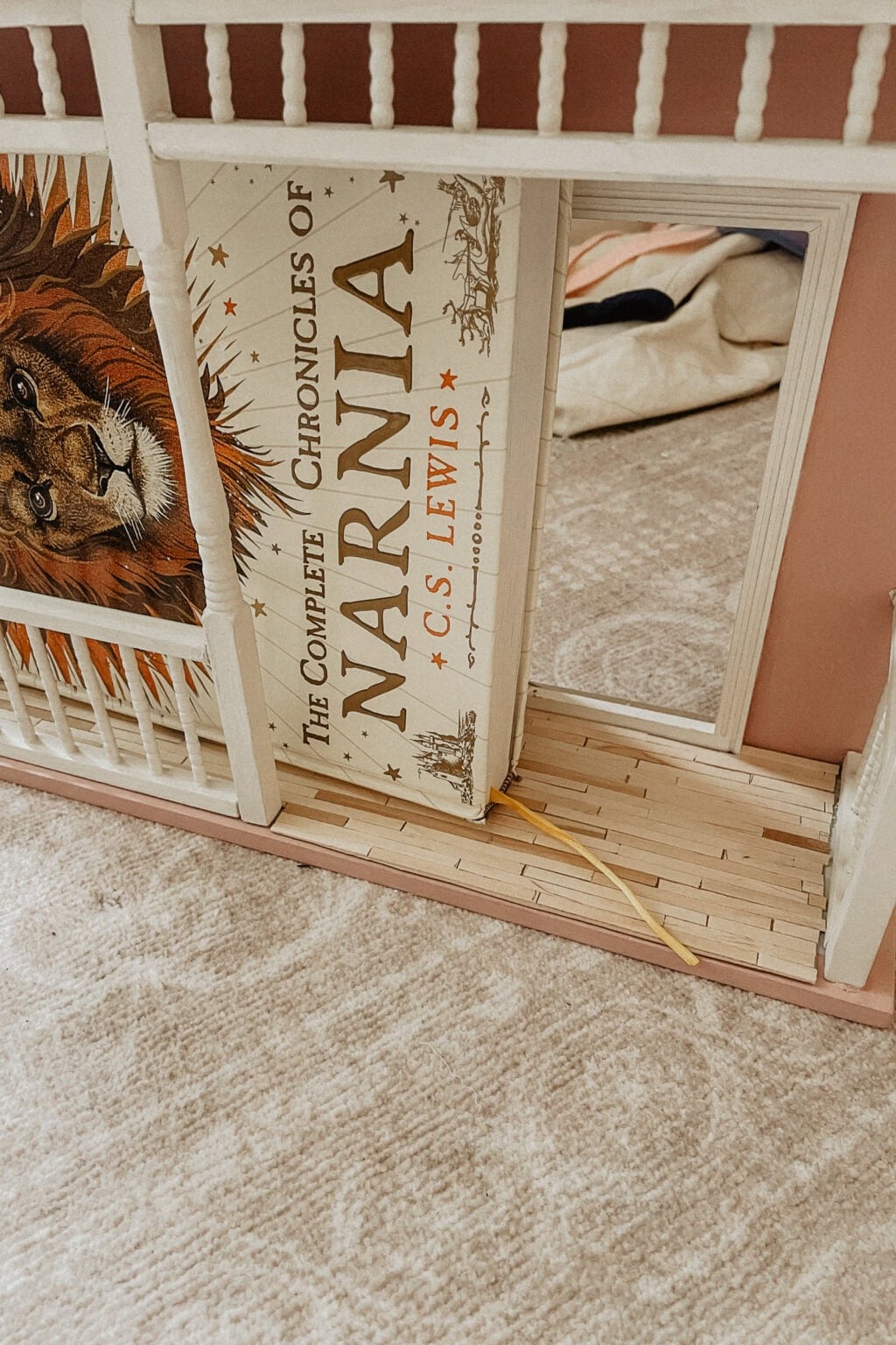 A large book titled The Complete Chronicles of Narnia by C.S. Lewis is propped up inside a miniature doll’s house, partially blocking the doorway and showing an illustration of a lion on the cover.