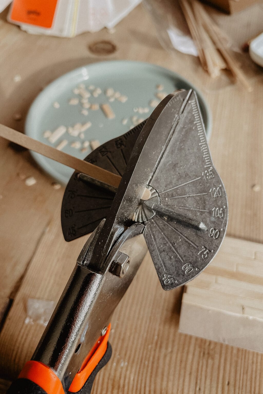 A close-up of a metal mitre shear tool cutting a thin wooden stick at an angle, with wood pieces and a green plate in the background on a wooden table.
