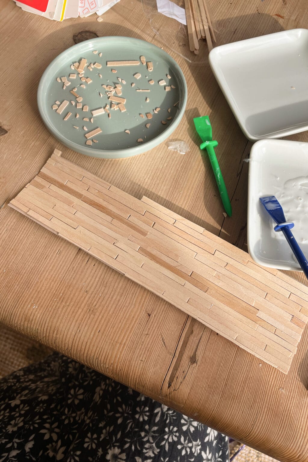 A wooden table with a plate of small wooden pieces, crafting tools, two white trays, and a partially assembled wood mosaic. The edge of a person’s patterned skirt is visible at the bottom.