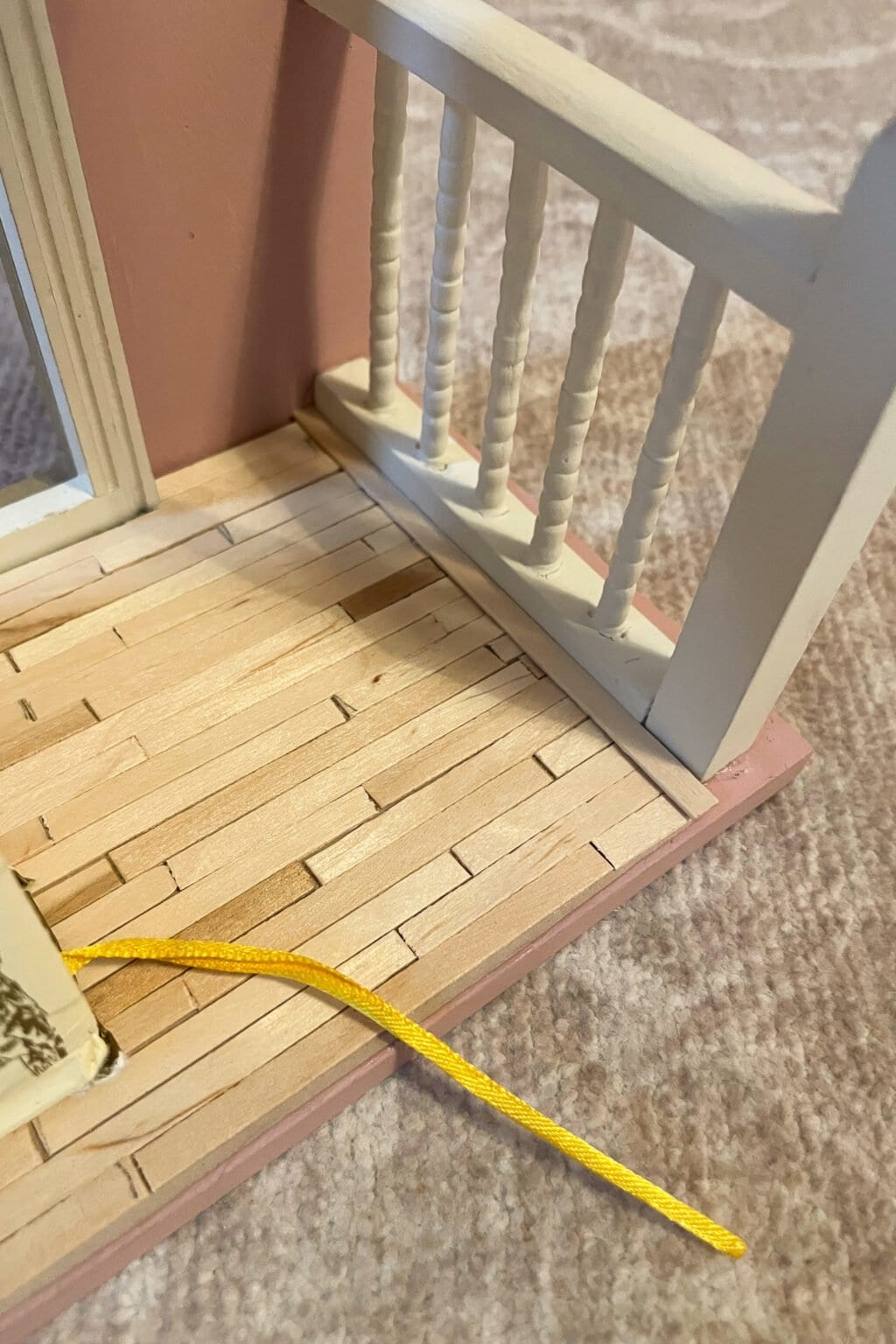 A yellow shoelace lies on the wooden floor of a doll’s house porch with white railings and a light pink wall in the background.