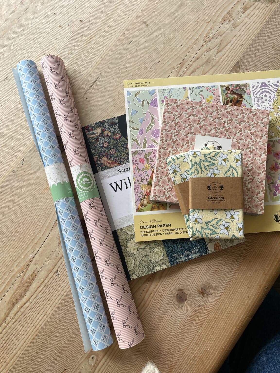 Two rolls of patterned wrapping paper and several packs of decorative paper with floral and geometric designs are arranged on a wooden table. The papers are in soft pastel and earth tones.