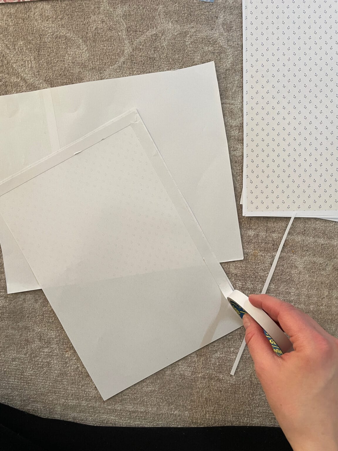 A hand holding a craft knife trims the edge of a sheet of white paper with a faint dot pattern, on a textured beige surface, with other sheets scattered nearby.