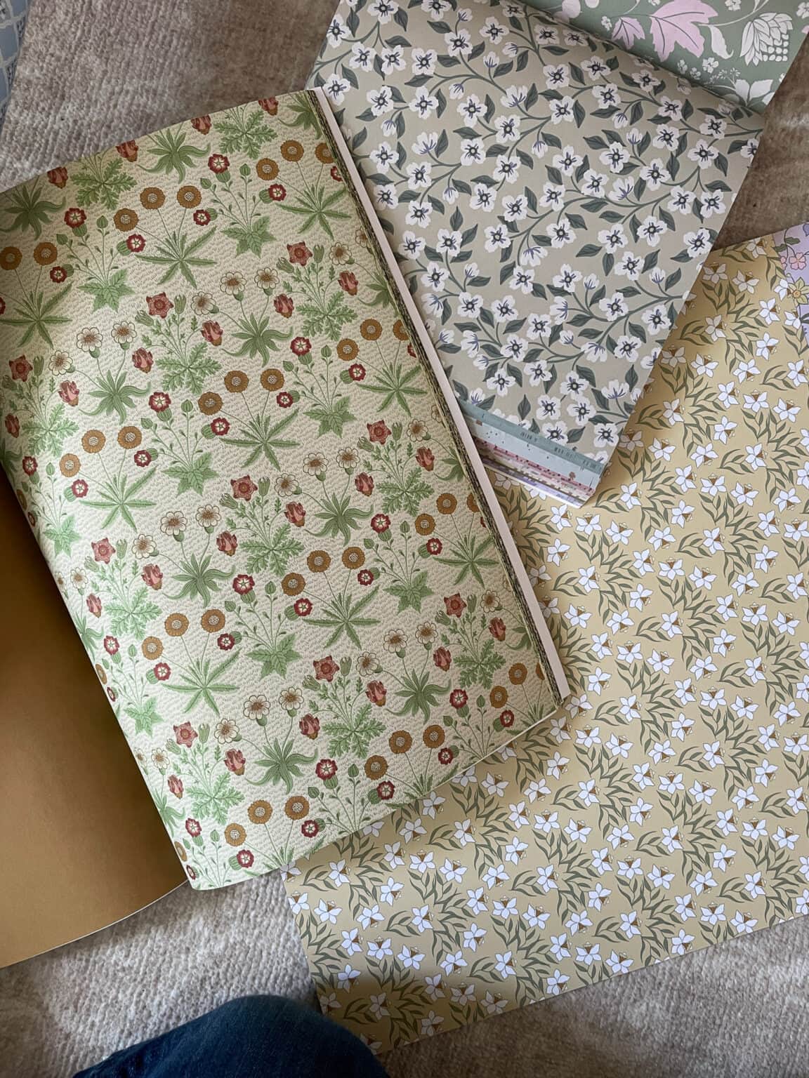 Three patterned wallpaper samples are laid out on a surface: one with green and brown floral designs, one with small white flowers on a yellow background, and one with white flowers and leaves on a grey background.