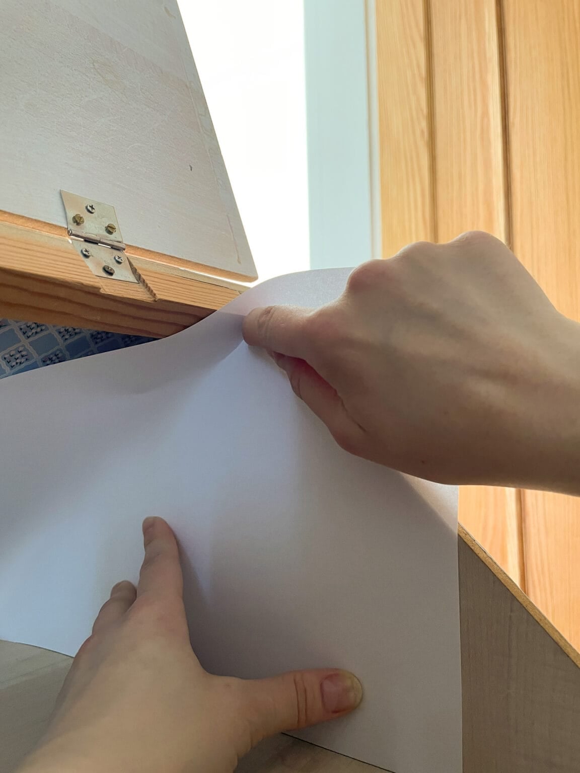 A person holds and smooths a white sheet of paper against the inside surface of a wooden box or drawer, next to a light wooden door.
