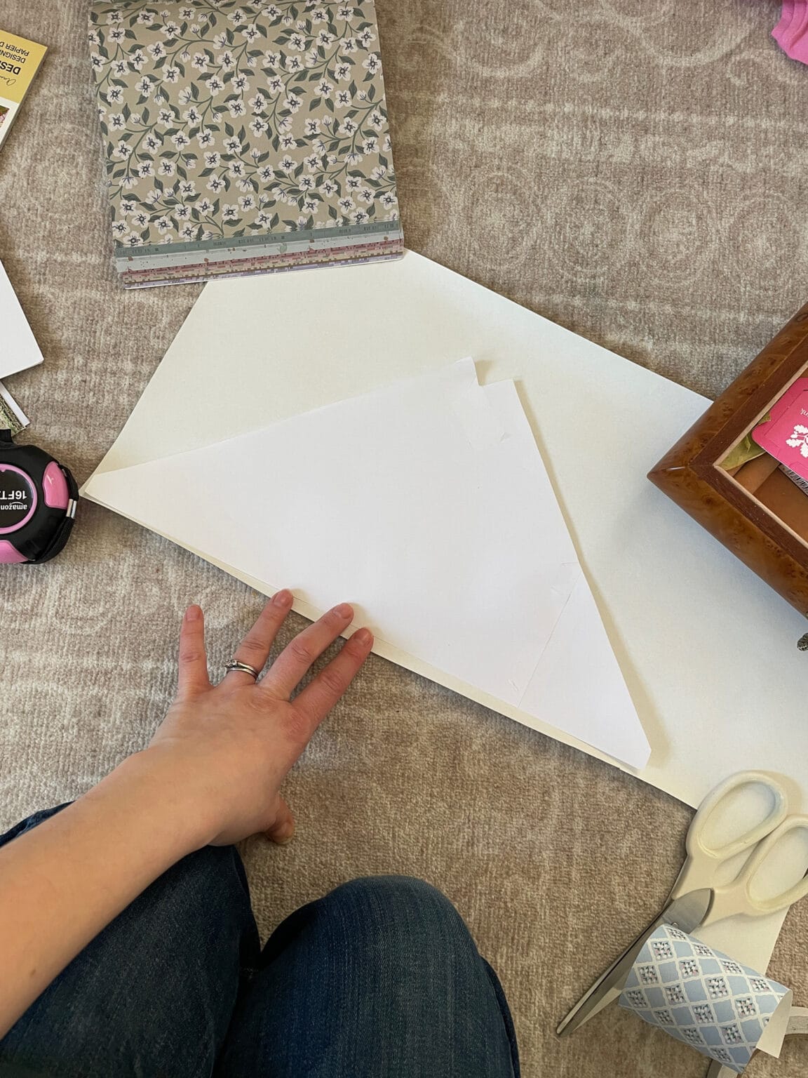 A person’s hand holds folded white paper on a cream-coloured patterned carpet, surrounded by scissors, a roll of tape, floral-patterned paper, a measuring tape, and part of a wooden box.